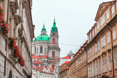 Aziz Nicholas Kilisesi, Prag Küçük Kasabası 'ndaki Barok Kilisesi, tarihi binalarla çerçevelenmiş yeşil kubbeleri olan dar bir caddenin üzerindeki kuleler. Pencere kutularındaki kırmızı çiçekler sahneye canlı bir dokunuş ekler