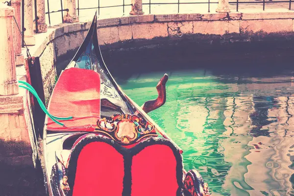 Taş bir duvar boyunca kenetlenmiş canlı kırmızı koltuklu geleneksel gondol, bir kanalın yeşilimsi suyuna yansıyor. Sahne, yumuşak ışıklandırmayla romantik bir Venedik atmosferini çağrıştıran klasik bir duyguya sahiptir.