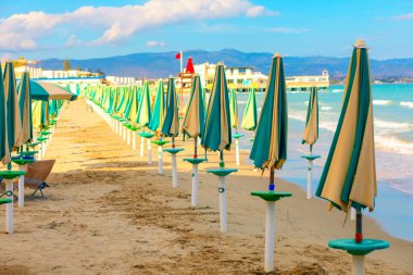 Kumsal, beyaz direklere asılmış kapalı yeşil ve bej renkli şemsiyelerle dolu. Arka planda binalar, kırmızı bayraklı bir iskele ve bulutlu bir gökyüzünün altında uzak dağlar var.