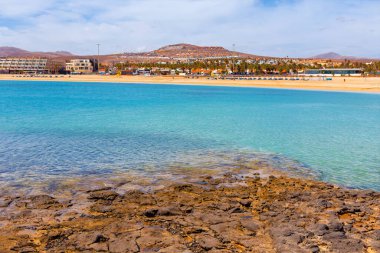 Caleta de Fuste plajı ve Fuerteventura 'da tatil köyleri. Sahil manzarası berrak turkuaz su, insanlarla ve binalarla dolu kumlu bir sahil, ve ön planda engebeli volkanik kayalar.