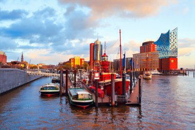 Hamburg limanına yanaşan tekneler, arka planda ikonik Elbphilharmonie yükselişiyle altın günbatımını yansıtıyor. Denizcilik cazibesiyle modern mimarinin dinamik bir karışımı.