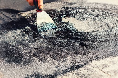 Çatı tamiri için kalın, siyah katranlı bir maddeyi kaba bir yüzeye serpmek için macun bıçağı kullanan bir adam. Maddenin parlak, ıslak bir görüntüsü ve küçük yansıtıcı parçacıkları var.