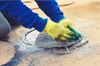 Bir insan diz çöküp bir çatı malzemesini katranlı bir maddeyle çevrili rögar kapağına bastırıyor. Sahne, işçiyle birlikte tamir sürecinin bir parçasıdır. 