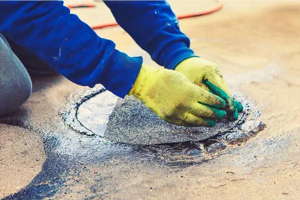 Bir insan diz çöküp bir çatı malzemesini katranlı bir maddeyle çevrili rögar kapağına bastırıyor. Sahne, işçiyle birlikte tamir sürecinin bir parçasıdır. 