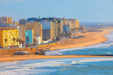 İspanya, Cadiz 'in kıyı manzarası, renkli binalarla çevrili uzun kumlu plaj, palmiye ağaçları gezinti güvertesi. Atlantik Okyanusunun gök mavisi dalgaları gökyüzünün altında yavaşça kıyıya çarpar.