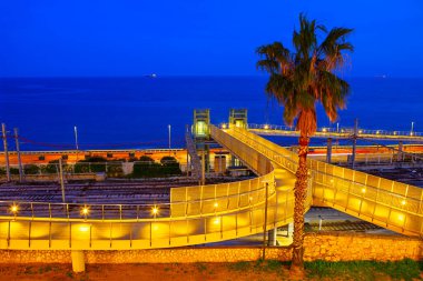 Tarragona 'daki gece sahnesi, kıyı şeridi yakınlarındaki demiryolu raylarının üzerinden geçen modern aydınlanmış yaya köprüsü ve palmiye ağaçları. Akdeniz karanlık bir gökyüzünün altında ufka doğru uzanıyor.