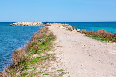 Sahil yolu, arkaplanı turkuaz su olan küçük bir limana doğru ilerliyor. Resimli sahil geçidi, karadan denize geçişi gösteriyor.