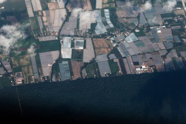 Aerial view of patchwork of agricultural fields, with numerous greenhouses and patches of cultivated land interspersed with small buildings. Landscape is shadowed by clouds and large dark area