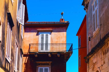 Beyaz kepenkleri ve işlenmiş demir balkonları olan bir Avrupa kasabasının mimari detayları. Dar bir caddede açık mavi gökyüzüne karşı renkli binalar.