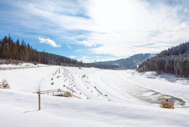Yoğun bir ormanın ortasında donmuş bir kış gölü. Güneş ışığı karla kaplı vadiyi aydınlatıyor, sakin kış sahnesini aydınlatıyor.