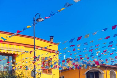 Renkli üçgen flama gölgeleri güneş ışığıyla aydınlatılmış cadde boyunca uzanır ve aşağıda neşeli gölgeler oluşturur. Şen şakrak cepheler ve sokak lambaları yaz aylarında, Latince esinlenilmiş mekanlarda ipucu veriyor.