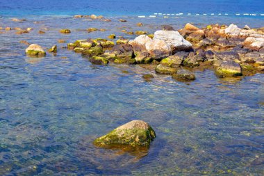 Deniz suyuna batırılmış taşlarla dolu sakin bir kıyı şeridi. Suyun yüzeyinin gökyüzünü yansıttığı sakin bir sahil manzarası. Kayalık su ve taşın etkileşimini gözler önüne seriyor