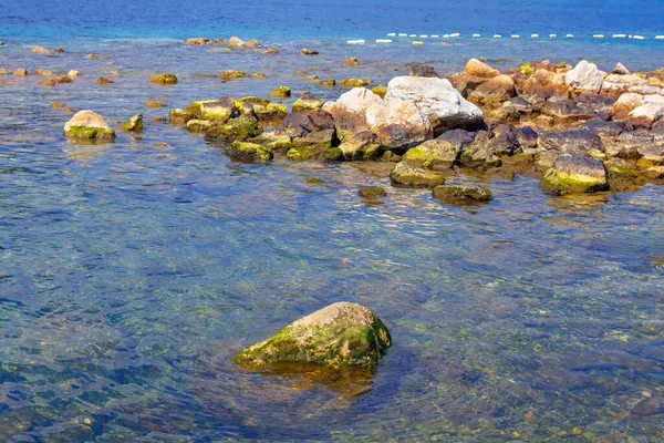 Deniz suyuna batırılmış taşlarla dolu sakin bir kıyı şeridi. Suyun yüzeyinin gökyüzünü yansıttığı sakin bir sahil manzarası. Kayalık su ve taşın etkileşimini gözler önüne seriyor