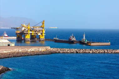 Endüstriyel bir limana yanaşan büyük bir açık deniz petrol platformu, sakin sularda römorkörlere yardım ediyor. Engebeli kıyı şeridi ve uzak dağlar deniz endüstrisinin bu sahnesini tamamlıyor.