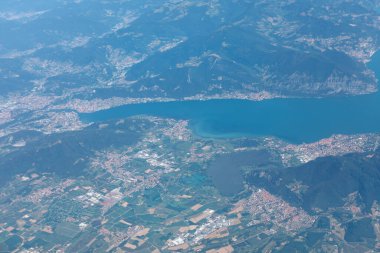 İtalyan Alpleri 'nde yeşil tarlalar ve engebeli dağlarla çevrili sakin göl manzarası. Kasabalar, doğal güzellikleri insan yerleşimleriyle harmanlayarak derin mavi suların etrafındaki manzarayı noktalar.