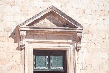 Süslü taş işçiliğine sahip üçgen bir soy ağacı yıpranmış bir taş cepheye açılan yeşil bir pencereyi taçlandırıyor. Klasik tasarım, eski mimariye zarafet ve simetri ekler.