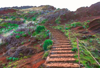 Taş basamaklar yeşil bitki örtüleri ve kırmızımsı topraklarla engebeli bir yamaçtan yükseliyor. Halat korkulukları yolu çiziyor, parlak bir gökyüzünün altındaki kayalık zirveye doğru gidiyor.