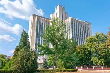 Chisinau 'daki görkemli hükümet binası çitlerin arkasında, dikey sütunlarla işaretlenmiş simetrik cephe ve yukarıda dalgalanan bir Moldova bayrağı şeklinde duruyor. Sahne, toplumsal formalite ve milli gurur yayar.