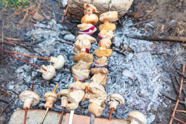 Mantar ve soğan şişleri açık hava kamp ateşinde sıcak küllerin üzerinde kızarıyor. Kırsal yemek sahnesi, doğada taze sebze hazırlamanın basitliğini yakalar.