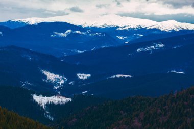 Karla kaplı zirveler orman tepelerinin ötesine yükseliyor, sislerle ve gölgelerle kaplı. Bulutlu gökyüzü engin, engebeli dağ manzarasının üzerine kasvetli bir hava salıyor.