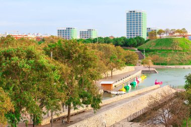 Valencia 'daki Cabecera Parkı ve göl, kuğu şekilleri de dahil olmak üzere canlı kürek tekneleriyle bezenmiş, yemyeşil ağaçlar ve belirgin çimenli bir tepe ile çevrili. Modern şehir binaları arka planda duruyor