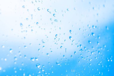 Numerous clear water droplets clinging to glass surface, set against softly blurred background of bright blue and white sky. Light refracts through the drops, creating a sense of freshness and calm