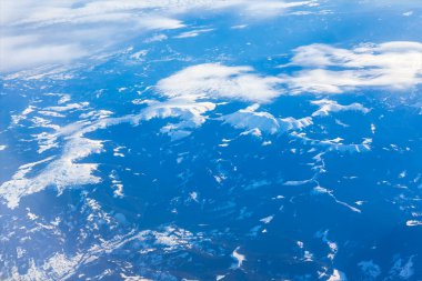 Kar dağlarının zirveleriyle kaplı, derinlik ve hareket katan sürüklenen bulutlarla kısmen örtülü. Yüksek irtifa görüntüsü dramatik tepeler ve zıt kayalık araziler gösterir