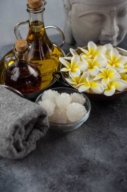 Spa massage relax composition setting. Natural essence oils, sea salt, towel, candles, Buddha head statue and plumeria frangipani exotic flowers. Gray colors.