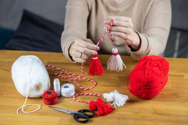 Woman making handmade traditional martisor, from red and white strings with tassel. Symbol of holiday 1 March, Martenitsa, Baba Marta, beginning of spring in Romania, Bulgaria, Moldova