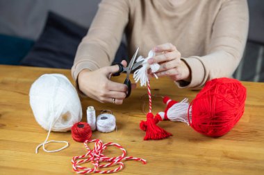 Woman making handmade traditional martisor, from red and white strings with tassel. Symbol of holiday 1 March, Martenitsa, Baba Marta, beginning of spring in Romania, Bulgaria, Moldova