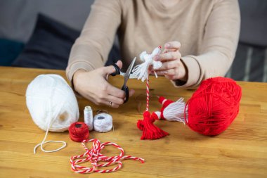 Woman making handmade traditional martisor, from red and white strings with tassel. Symbol of holiday 1 March, Martenitsa, Baba Marta, beginning of spring in Romania, Bulgaria, Moldova