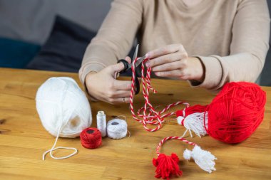 Woman making handmade traditional martisor, from red and white strings with tassel. Symbol of holiday 1 March, Martenitsa, Baba Marta, beginning of spring in Romania, Bulgaria, Moldova