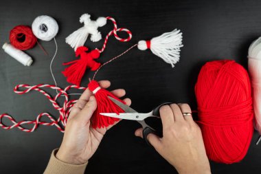 Woman making handmade traditional martisor, from red and white strings with tassel. Symbol of holiday 1 March, Martenitsa, Baba Marta, beginning of spring in Romania, Bulgaria, Moldova