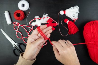 Woman making handmade traditional martisor, from red and white strings with tassel. Symbol of holiday 1 March, Martenitsa, Baba Marta, beginning of spring in Romania, Bulgaria, Moldova