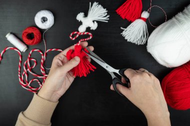 Woman making handmade traditional martisor, from red and white strings with tassel. Symbol of holiday 1 March, Martenitsa, Baba Marta, beginning of spring in Romania, Bulgaria, Moldova