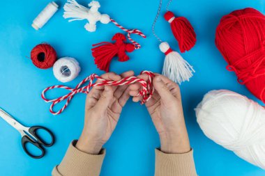 Woman making handmade traditional martisor, from red and white strings with tassel. Symbol of holiday 1 March, Martenitsa, Baba Marta, beginning of spring in Romania, Bulgaria, Moldova