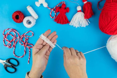 Woman making handmade traditional martisor, from red and white strings with tassel. Symbol of holiday 1 March, Martenitsa, Baba Marta, beginning of spring in Romania, Bulgaria, Moldova