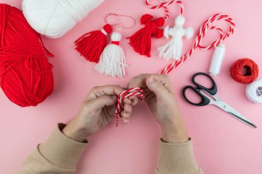 Woman making handmade traditional martisor, from red and white strings with tassel. Symbol of holiday 1 March, Martenitsa, Baba Marta, beginning of spring in Romania, Bulgaria, Moldova