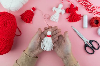 Woman making handmade traditional martisor, from red and white strings with tassel. Symbol of holiday 1 March, Martenitsa, Baba Marta, beginning of spring in Romania, Bulgaria, Moldova clipart