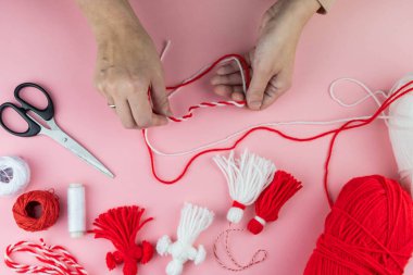 Woman making handmade traditional martisor, from red and white strings with tassel. Symbol of holiday 1 March, Martenitsa, Baba Marta, beginning of spring in Romania, Bulgaria, Moldova