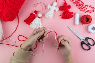 Woman making handmade traditional martisor, from red and white strings with tassel. Symbol of holiday 1 March, Martenitsa, Baba Marta, beginning of spring in Romania, Bulgaria, Moldova