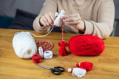 Woman making handmade traditional martisor, from red and white strings with tassel. Symbol of holiday 1 March, Martenitsa, Baba Marta, beginning of spring in Romania, Bulgaria, Moldova