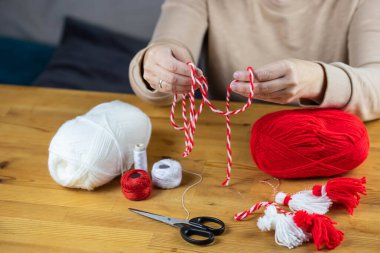 Woman making handmade traditional martisor, from red and white strings with tassel. Symbol of holiday 1 March, Martenitsa, Baba Marta, beginning of spring in Romania, Bulgaria, Moldova