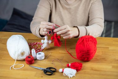 Woman making handmade traditional martisor, from red and white strings with tassel. Symbol of holiday 1 March, Martenitsa, Baba Marta, beginning of spring in Romania, Bulgaria, Moldova