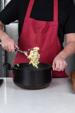 Adam şef, kırmızı önlükle giyilmiş, ustalıkla haşlanmış patatesleri tencerede eziyor, pürüzsüz, ev yapımı patates püresi yapıyor. Sahne, aşçılık ustalığını ve rahatlatıcı bir yemek hazırlama zevkini yansıtıyor..