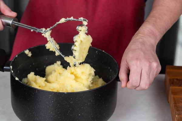 Man chef, donned in a red apron, skillfully mashes boiled potatoes in a pot, creating smooth, homemade mashed potato. The scene radiates culinary mastery and the joy of crafting a comforting dish.