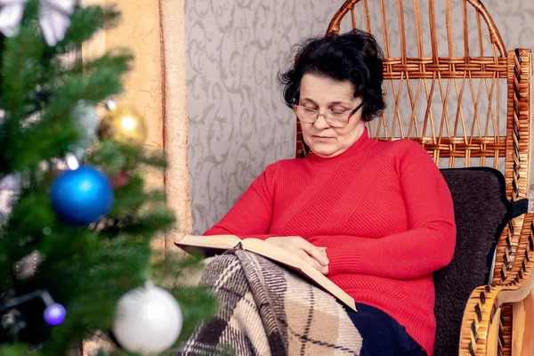 An elderly woman reads a book, the Bible, in a wicker chair in front of a Christmas tree. Celebrating Christmas alone