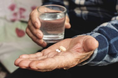 Yaşlı bir adam elinde haplar ve bir bardak su tutuyor. Hastalığı tedavi etmek için hap alıyorum.