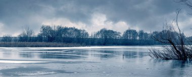 Nehirli kış manzarası, sudaki ağaçların yansıması ve karanlık tonlarda kasvetli havada dramatik bulutlu gökyüzü.