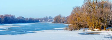 Güneşli bir günde, buzla kaplı bir nehrin yakınındaki ağaçların olduğu kış manzarası.